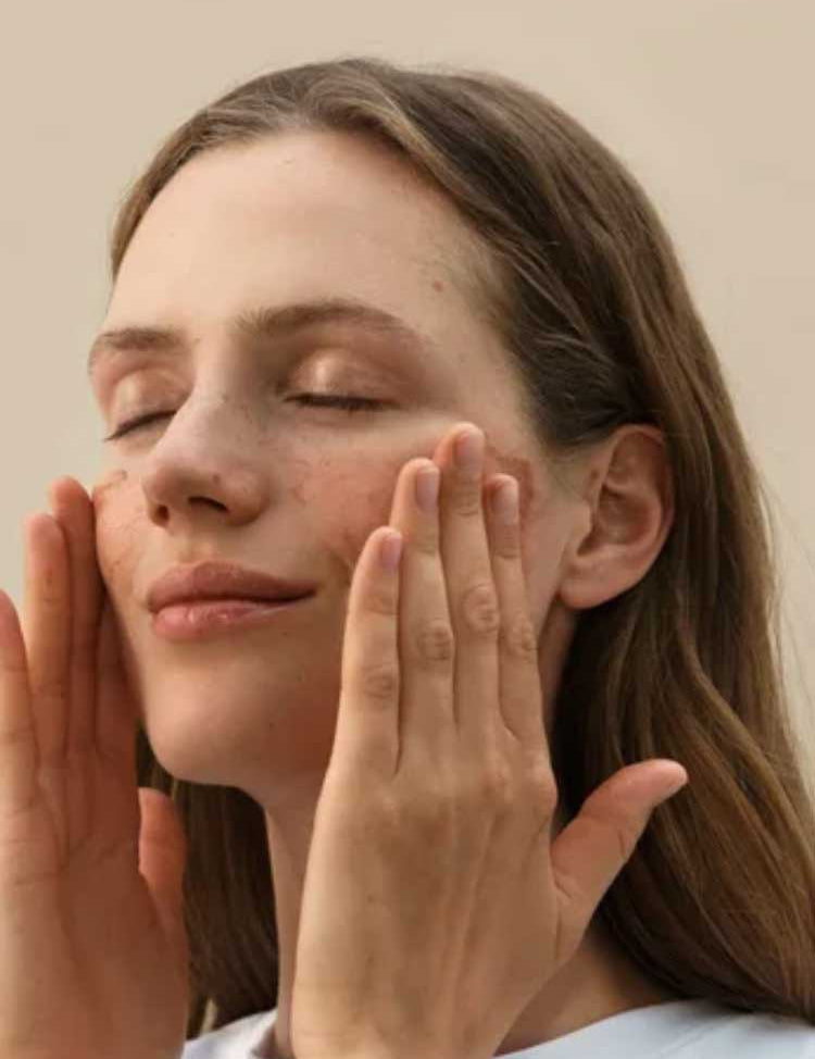 A woman with long brown hair smiles softly, gently touching her cheeks as her glowing skin radiates health—showcasing the effect of Dr Hauschka Translucent Bronzing Tint 18ml for a naturally radiant look.