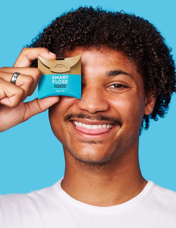 Smiling curly-haired man holding a tan and turquoise Dr Tung's Activated Charcoal Smart Floss dispenser over one eye against a turquoise background.
