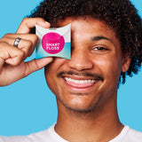 Smiling curly-haired man holding a white Dr Tung's Smart Floss dispenser with a bright pink circle over one eye against a turquoise background.