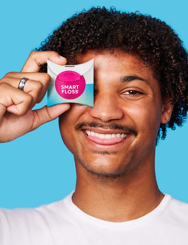 Smiling curly-haired man holding a white Dr Tung's Smart Floss dispenser with a bright pink circle over one eye against a turquoise background.