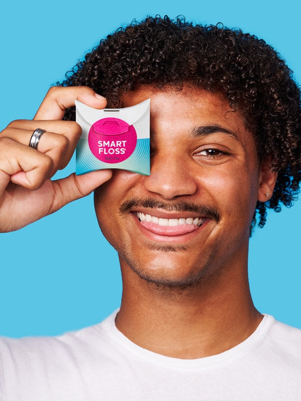 Smiling curly-haired man holding a white Dr Tung's Smart Floss dispenser with a bright pink circle over one eye against a turquoise background.