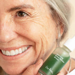 Smiling woman with grey hair holding the Ere Perez Quandong Green Booster Serum bottle beside her cheek