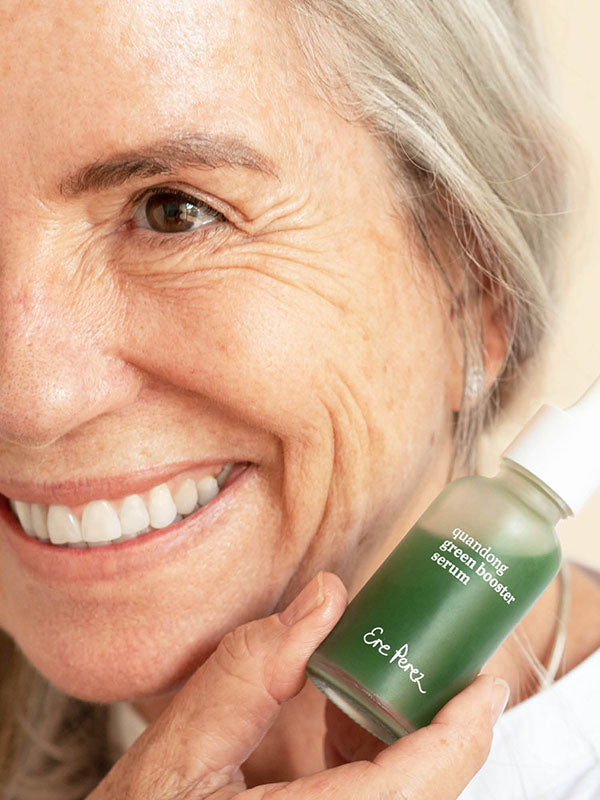 Smiling woman with grey hair holding the Ere Perez Quandong Green Booster Serum bottle beside her cheek