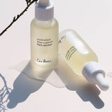 Two frosted bottles of Ere Perez Australian Blue Cypress Face Nectar with a cypress sprig on a cream background