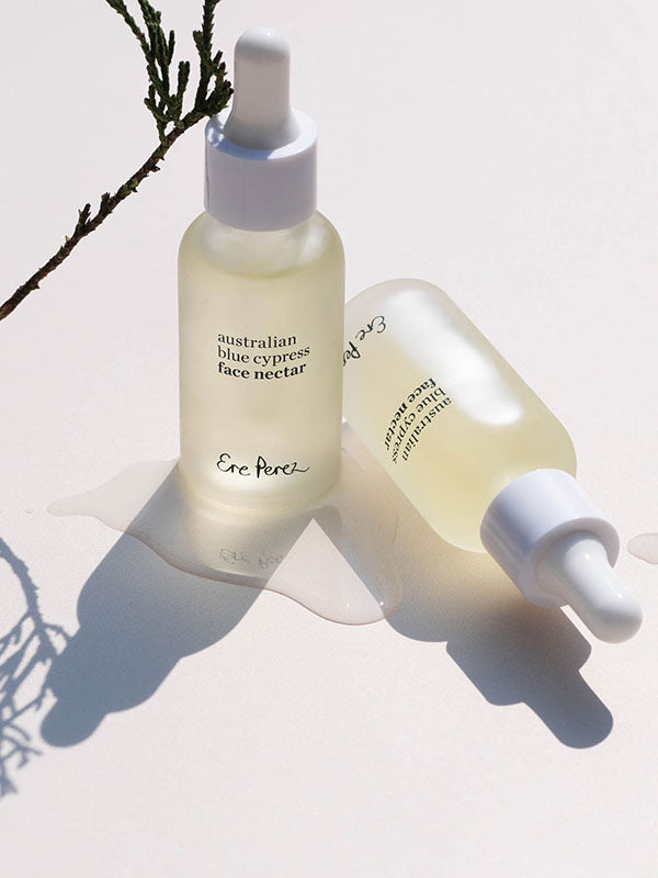 Two frosted bottles of Ere Perez Australian Blue Cypress Face Nectar with a cypress sprig on a cream background