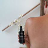 Woman holding the Mukti Rose Blossom Hydrating Mist over her bare shoulder with flannel flowers