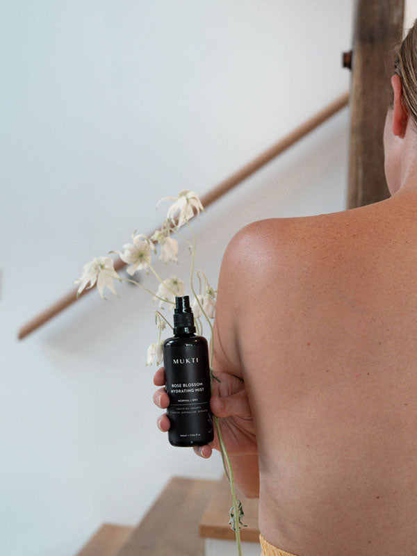 Woman holding the Mukti Rose Blossom Hydrating Mist over her bare shoulder with flannel flowers