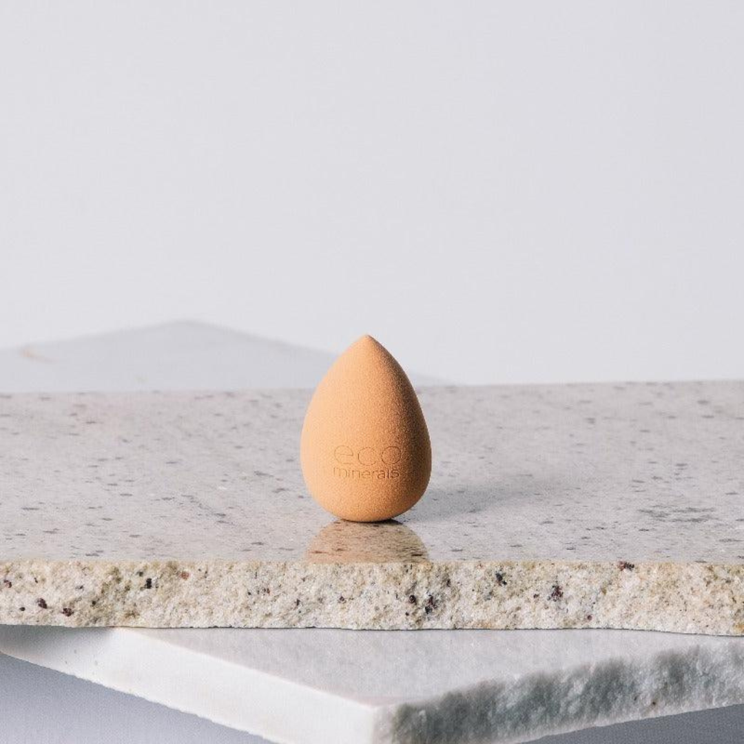 Peach-coloured, teardrop-shaped Eco Minerals Beauty Blender standing upright on a rough pale stone slab, with a pointed top and rounded base.