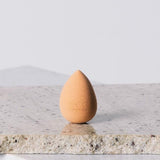 Peach-coloured, teardrop-shaped Eco Minerals Beauty Blender standing upright on a rough pale stone slab, with a pointed top and rounded base.
