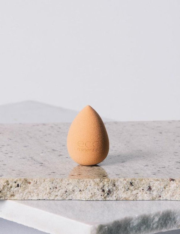 Peach-coloured, teardrop-shaped Eco Minerals Beauty Blender standing upright on a rough pale stone slab, with a pointed top and rounded base.