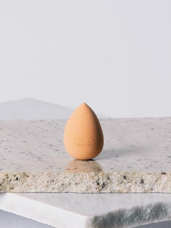 Peach-coloured, teardrop-shaped Eco Minerals Beauty Blender standing upright on a rough pale stone slab, with a pointed top and rounded base.