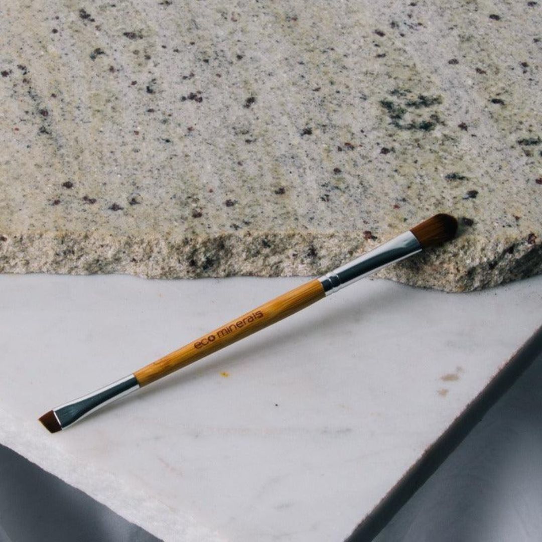 Eco Minerals Duo Brush lying diagonally across pale stone slabs, with a bamboo handle, silver ferrules, a rounded brown brush and a small angled brush at opposite ends.