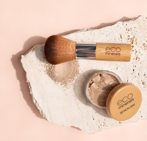 Open Eco Minerals Flawless Matte Foundation pot with a tan lid beside a bamboo-handled brush and spilled beige powder on a rough white stone slab against peach.