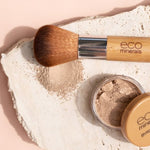 Open Eco Minerals Flawless Matte Foundation pot with a tan lid beside a bamboo-handled brush and spilled beige powder on a rough white stone slab against peach.