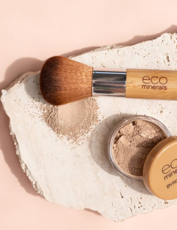 Open Eco Minerals Flawless Matte Foundation pot with a tan lid beside a bamboo-handled brush and spilled beige powder on a rough white stone slab against peach.
