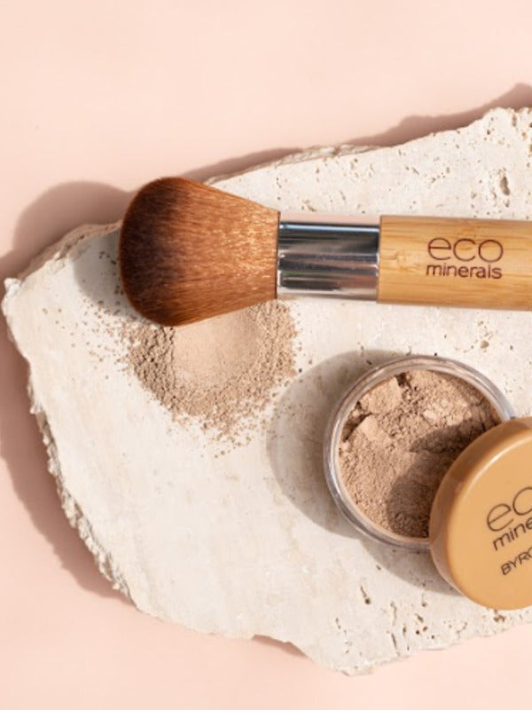 Open Eco Minerals Flawless Matte Foundation pot with a tan lid beside a bamboo-handled brush and spilled beige powder on a rough white stone slab against peach.