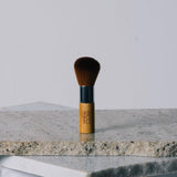 Eco Minerals Kabuki Brush standing upright on a pale stone slab, with rounded brown bristles, a silver ferrule and a short bamboo handle.