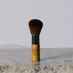 Eco Minerals Kabuki Brush standing upright on a pale stone slab, with rounded brown bristles, a silver ferrule and a short bamboo handle.