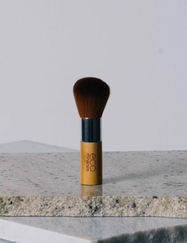 Eco Minerals Kabuki Brush standing upright on a pale stone slab, with rounded brown bristles, a silver ferrule and a short bamboo handle.