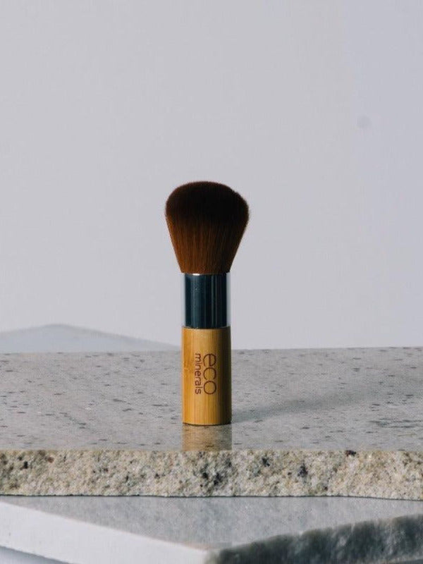 Eco Minerals Kabuki Brush standing upright on a pale stone slab, with rounded brown bristles, a silver ferrule and a short bamboo handle.