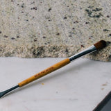 Eco Minerals Duo Brush lying diagonally across pale stone slabs, with a bamboo handle, silver ferrules, a rounded brown brush and a small angled brush at opposite ends.