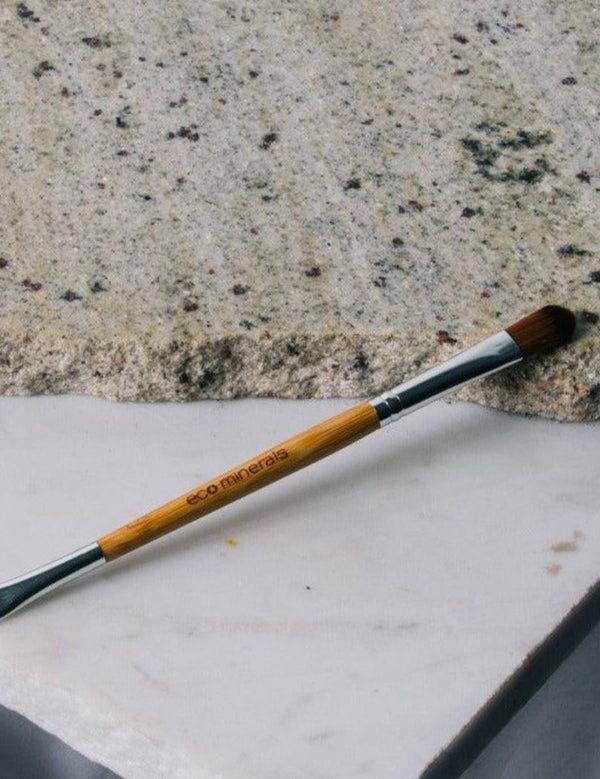 Eco Minerals Duo Brush lying diagonally across pale stone slabs, with a bamboo handle, silver ferrules, a rounded brown brush and a small angled brush at opposite ends.