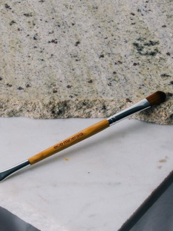 Eco Minerals Duo Brush lying diagonally across pale stone slabs, with a bamboo handle, silver ferrules, a rounded brown brush and a small angled brush at opposite ends.