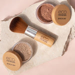 Two open Eco Minerals Perfection Dewy Mineral Foundation powder pots with a bamboo-handled brush on a textured white stone slab against peach.