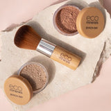 Two open Eco Minerals Perfection Dewy Mineral Foundation powder pots with a bamboo-handled brush on a textured white stone slab against peach.