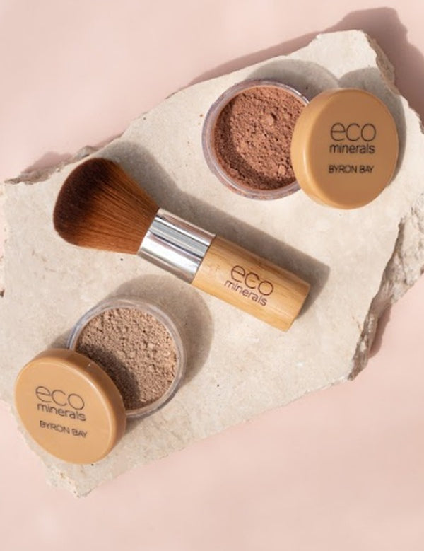 Two open Eco Minerals Perfection Dewy Mineral Foundation powder pots with a bamboo-handled brush on a textured white stone slab against peach.