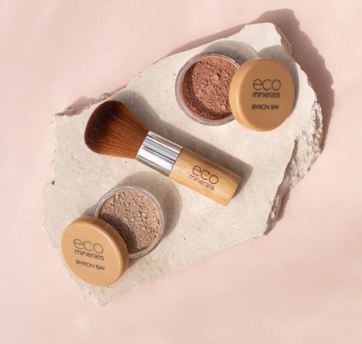 Two open Eco Minerals powder pots with pale beige and warm bronze powders, tan lids and a bamboo-handled kabuki brush on a rough white stone slab against peach.