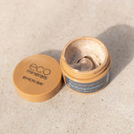 Open Eco Minerals Purifying Exfoliator jar showing a swirl of grainy beige cream, with its tan lid beside it on a pale stone surface.