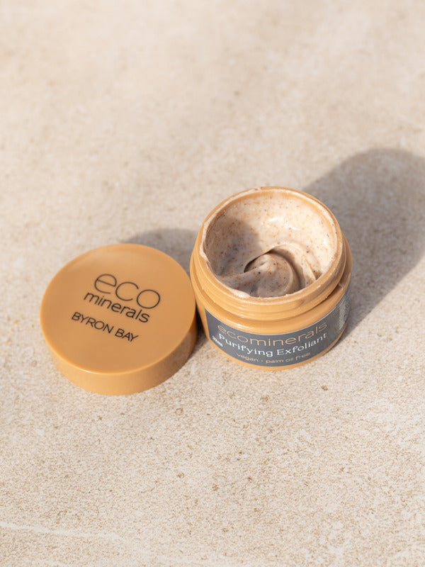 Open Eco Minerals Purifying Exfoliator jar showing a swirl of grainy beige cream, with its tan lid beside it on a pale stone surface.
