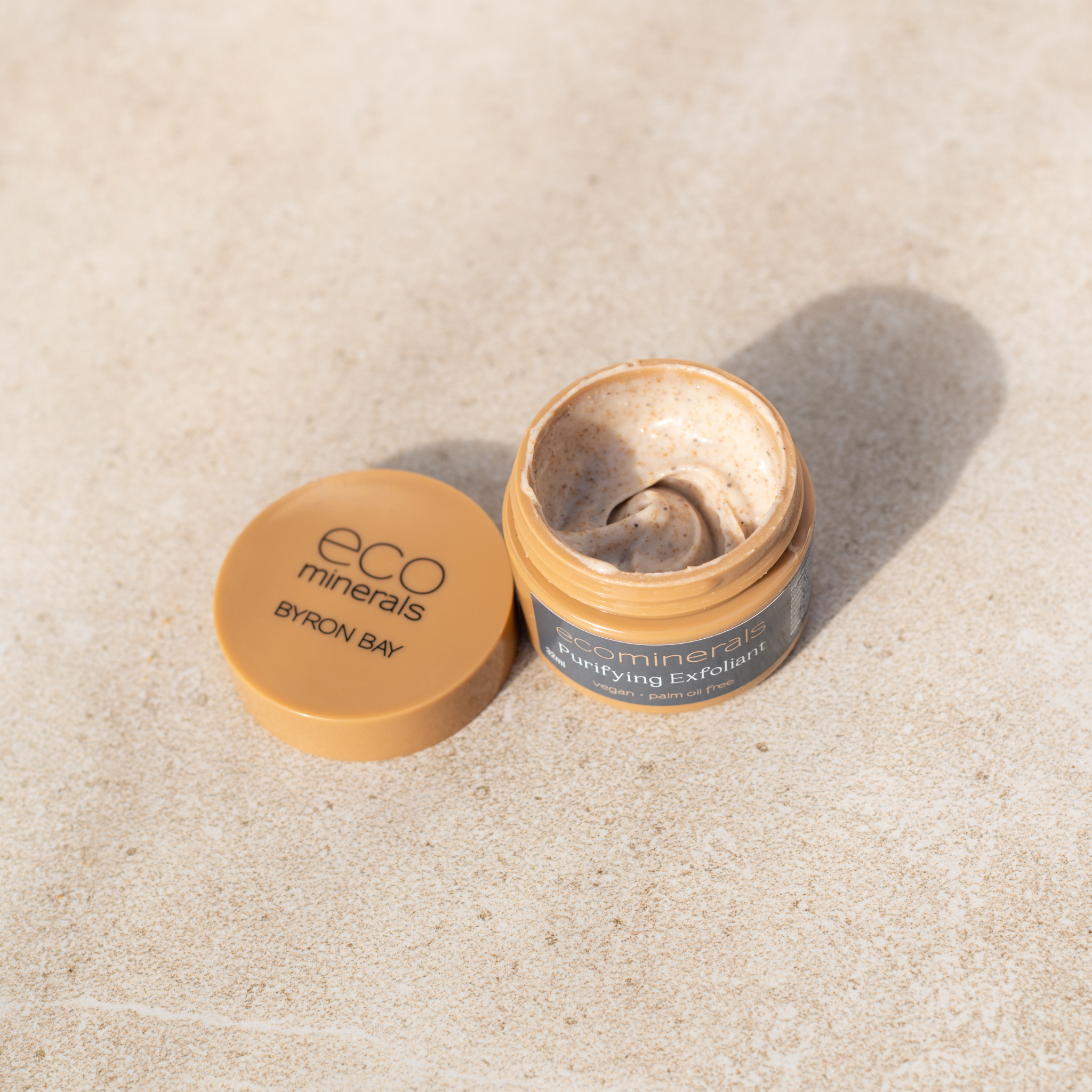 Open Eco Minerals Purifying Exfoliator jar showing a swirl of grainy beige cream, with its tan lid beside it on a pale stone surface.