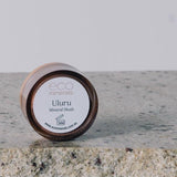 Round Eco Minerals Uluru blush pot resting on its edge on a pale stone slab, with a small pile of loose reddish-peach powder on the reflective surface below.
