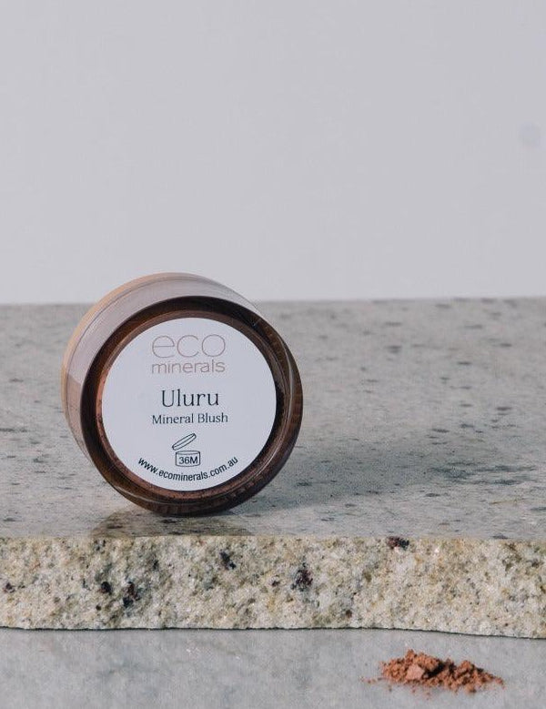 Round Eco Minerals Uluru blush pot resting on its edge on a pale stone slab, with a small pile of loose reddish-peach powder on the reflective surface below.