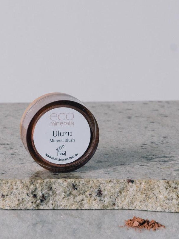 Round Eco Minerals Uluru blush pot resting on its edge on a pale stone slab, with a small pile of loose reddish-peach powder on the reflective surface below.