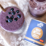 Overhead view of a purple smoothie topped with blueberries beside a glass jug and a blue Eden Healthfoods High Octane tin on a pale cloth.