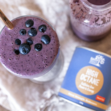 Overhead view of a purple smoothie topped with blueberries beside a glass jug and a blue Eden Healthfoods High Octane tin on a pale cloth.