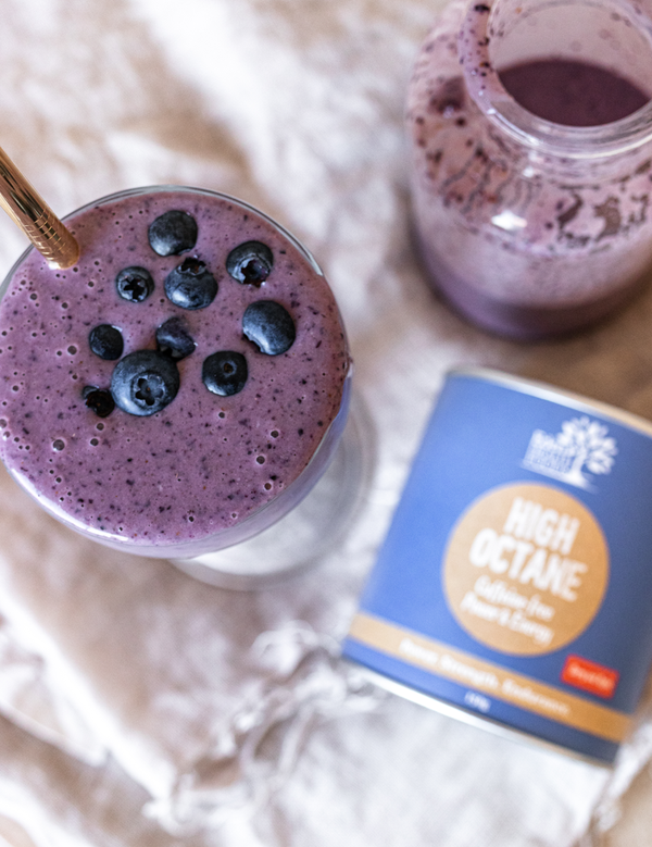 Overhead view of a purple smoothie topped with blueberries beside a glass jug and a blue Eden Healthfoods High Octane tin on a pale cloth.