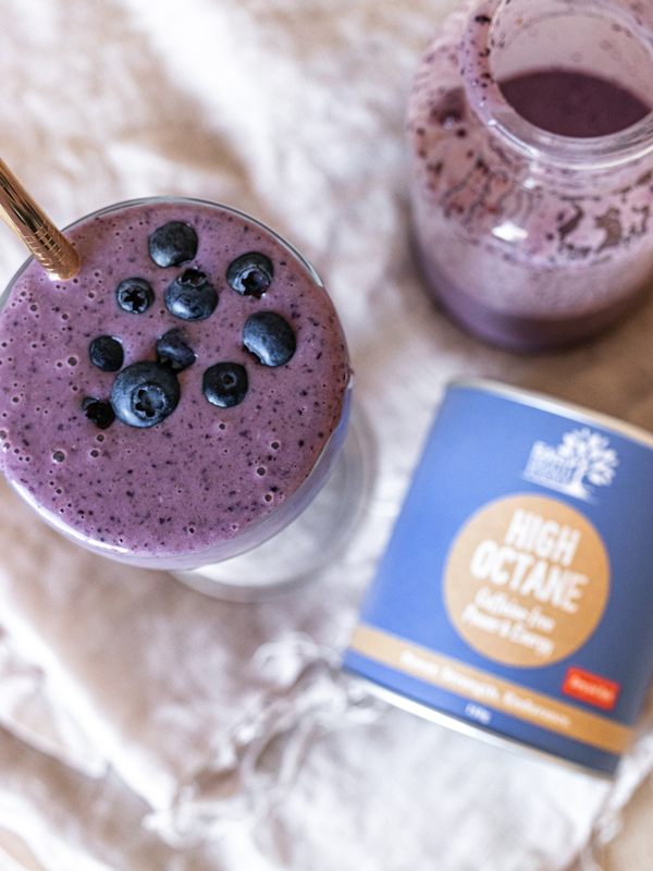 Overhead view of a purple smoothie topped with blueberries beside a glass jug and a blue Eden Healthfoods High Octane tin on a pale cloth.