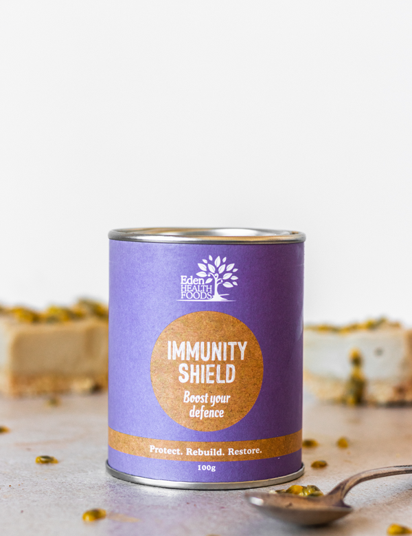 Purple Eden Healthfoods Immunity Shield tin on a pale surface beside a spoon, with tiny yellow flowers scattered around it and pale blocks behind.