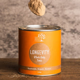 A spoon heaped with beige-brown powder held above an open orange Eden Healthfoods Longevity tin on a wooden table, with a pale stone wall behind.