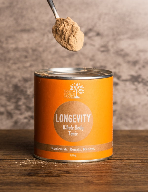 A spoon heaped with beige-brown powder held above an open orange Eden Healthfoods Longevity tin on a wooden table, with a pale stone wall behind.