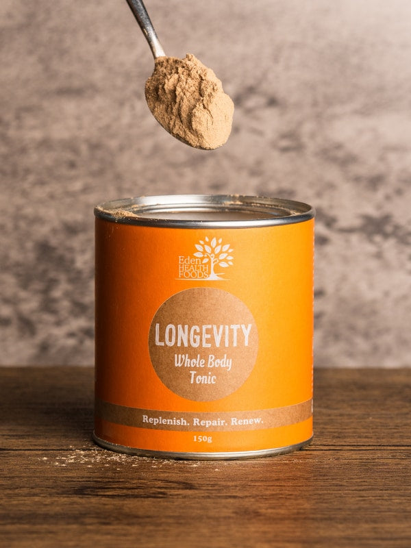 A spoon heaped with beige-brown powder held above an open orange Eden Healthfoods Longevity tin on a wooden table, with a pale stone wall behind.