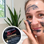 A woman spreads dark grey Elektra Charcoal Pomegranate Cleanser onto her cheek with her fingertips; patches cover her forehead and chin, and the labelled lid is beside her.