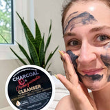 A woman spreads dark grey Elektra Charcoal Pomegranate Cleanser onto her cheek with her fingertips; patches cover her forehead and chin, and the labelled lid is beside her.