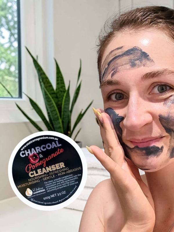 A woman spreads dark grey Elektra Charcoal Pomegranate Cleanser onto her cheek with her fingertips; patches cover her forehead and chin, and the labelled lid is beside her.