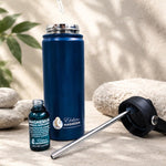 A hand holds a glass dropper above an open blue Elektra water bottle; a small magnesium supplement bottle and the detached lid with metal straw sit beside it.