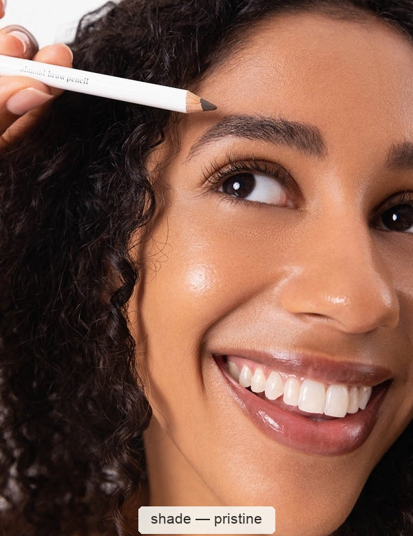 A smiling woman with dark curly hair applies a white Ere Perez Almond Brow Pencil to her eyebrow; the caption below names the shade Pristine.